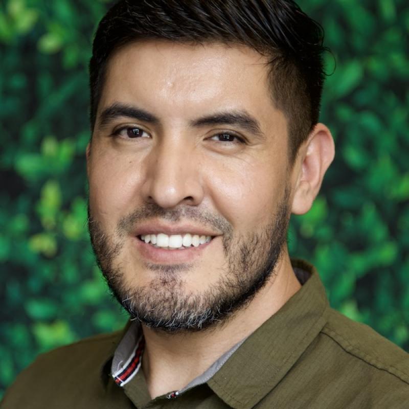 A photo of Enrique Campos wearing an earth green button-down shirt with a green background.