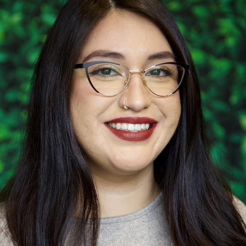 A photo of Zuleika Bravo wearing a gray shirt with a green background.